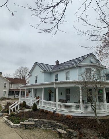 White two-story house with wraparound porch and metal roof on overcast day