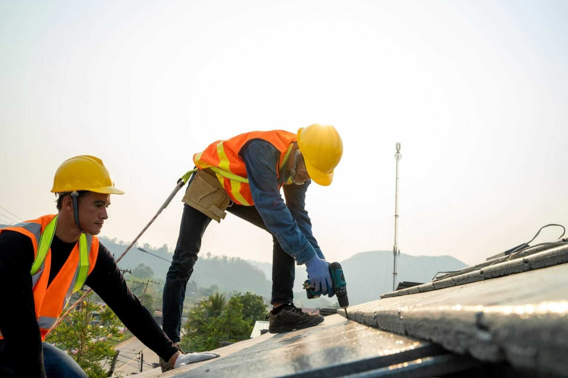 Two construction workers in safety gear drilling on concrete rooftop with mountains