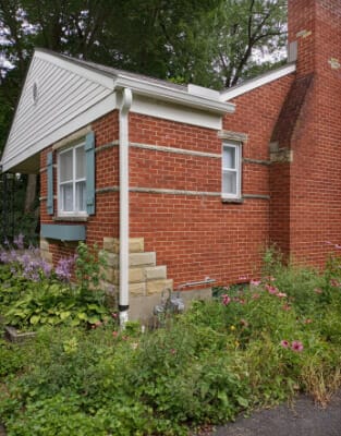 Red brick house with white trim and metal roof surrounded by lush green garden