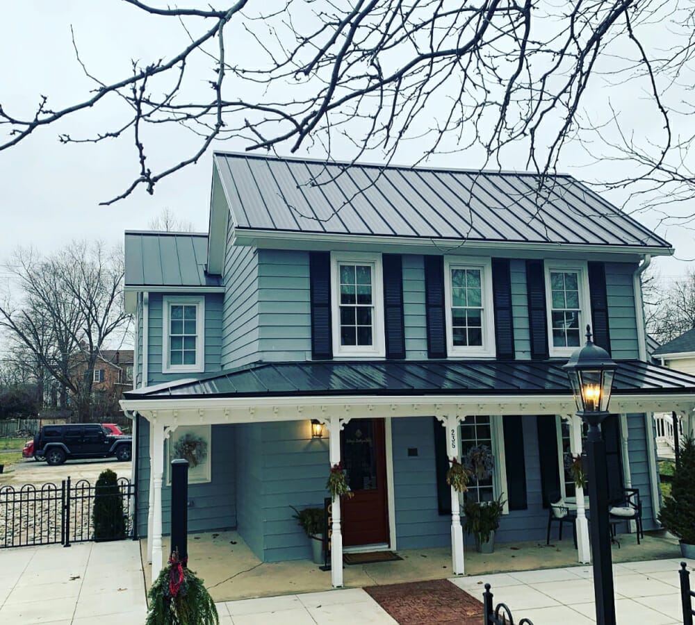 Two-story sage green house with black shutters and metal roof featuring front porch